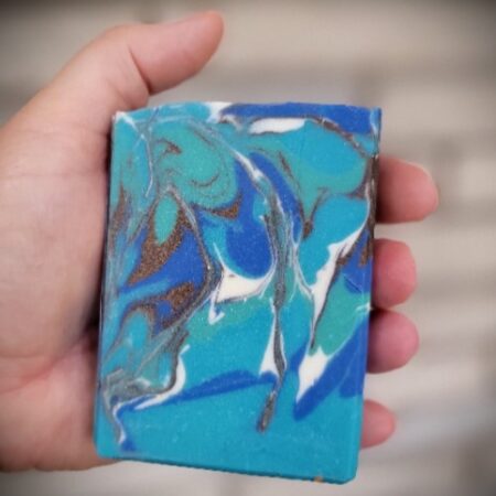 handmade soap in hand for size. Soap is swirled with colours of blue, teal, white and brown