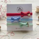 Set of three embroidered baby headbands (red, blue, mint) displayed on a wooden table with sewing threads and flowers.