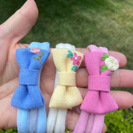 Three hand-embroidered baby headbands in pastel tones — blue, yellow, and pink — each featuring delicate floral embroidery, displayed outdoors in natural light.