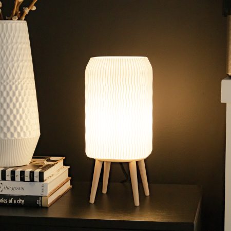 Tall modern desk lamp with a tall rectangular shade and minimalist beige base, emitting a warm LED glow. Placed on a wooden side table next to some plants and a fireplace, the lamp adds soft, functional light for work or reading. Perfect as a simple bedside lamp, cozy living room light, or housewarming gift for a modern home.