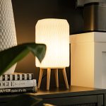 Tall modern desk lamp with a tall rectangular shade and minimalist beige base, emitting a warm LED glow. Placed on a wooden side table next to some plants and a fireplace, the lamp adds soft, functional light for work or reading. Perfect as a simple bedside lamp, cozy living room light, or housewarming gift for a modern home.